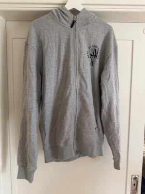 Grå zip hoodie från GASP - Snygg grå zip hoodie från GASP med tryck av en dödskalle och texten 'NO COMPROMISES' på bröstet. Hoodien har dragkedja, två fickor fram och en klassisk huva. Tillverkad i mjukt bomullsmaterial som är skönt att bära. Tröjan är helt ny och oanvänd