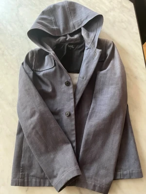 Grå hooded jacka från A.P.C. - Snygg grå jacka från A.P.C. med huva och knäppning framtill. Jackan har en minimalistisk design med raka linjer och långa ärmar. Tillverkad i ett slätt tyg som känns lätt och passar perfekt till vardagslooken.