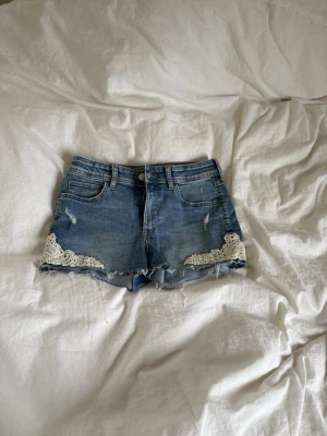 Blå Lågmidjade jeansshorts med spetsdetaljer - Säljer ett par blå jeansshorts med råa kanter och vita spetsdetaljer vid bensluten. Shortsen har basic fickor och är justerbara i midjan. Dom är mer Lågmidjade än dom ser ut på bilden. Inga fläckar eller slitningar. 