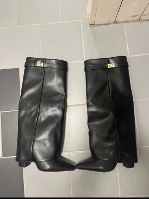 Svarta höga boots med klack - Snygga svarta boots i slätt skinnimitation med bred, hög skaft och markerad klack. Dekorativt spänne upptill och dragkedja på insidan. Perfekta för att lyfta din outfit med en edgy vibe.