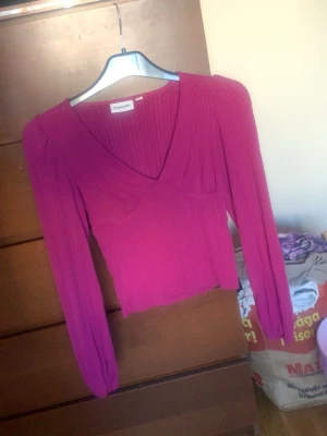 Plisserad rosa topp från Kappahl  - Trendig rosa topp från kappahl med plisserat tyg och v-ringad hals. Toppen har långa ärmar och en figurnära passform som ger en snygg siluett. Perfekt att styla med jeans eller kjol för en cool look.
