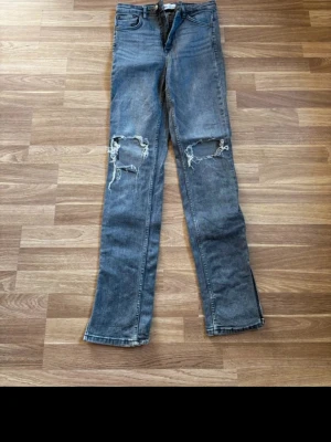 Grå slitna raka jeans med hål - Säljer ett par grå raka jeans med stora slitna hål på båda knäna. Jeansen har klassisk femficksdesign och är tillverkade i denim. Perfekta för en avslappnad och trendig streetstyle-look.