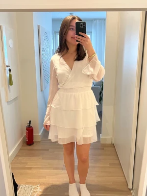 Vit studentklänning - Söt studentklänning 👩‍🎓Aldrig använd (lappen sitter kvar) Kortklänning med omlottliv och v-ringning. Klänningen har långa, transparenta ärmar och flera lager volanger i kjolen. 