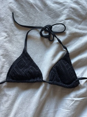 Svart trekantsbikini med struktur - Snygg svart trekantsbikini med knyt i nacken och ryggen. Bikinin har en strukturerad, nästan virkad look som ger en trendig känsla. Perfekt för lata dagar på stranden eller poolhäng med vänner.