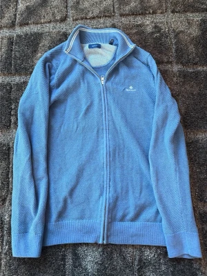 Blå ziptröja från GANT - Snygg blå ziptröja från GANT med diskret logga på bröstet. Tröjan har ribbade muddar, hög krage med vit detalj och är stickad i ett mjukt material. Perfekt för lager-på-lager och ger en clean look. OBS ETT HÅL PÅ ARMEN DÄR AV PRISET!