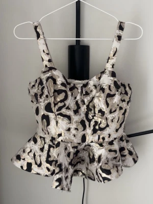 Leopardmönstrad korsettopp med volang - Snygg korsettopp med leopardmönster i svart, beige och guld. Toppen har breda axelband, markerade kupor och volang nedtill. Dragkedja bak för enkel påklädning. De guldfärgade delarna glänser fint i solen. Perfekt för dig som vill sticka ut med en trendig look. Storlek M men motsvarar snarare S eller XS. Ursprungligt pris 100 €.