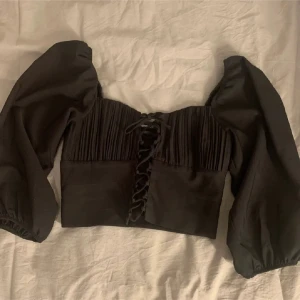 Svart topp med puffärmar Gina - Trendig svart topp med puffärmar och snörning framtill. Toppen har plisserad detalj vid bysten och är tillverkad i ett mjukt tyg som ger en snygg passform. Perfekt för dig som vill sticka ut med en cool och stilren look. Strl 32 men funkar som 34