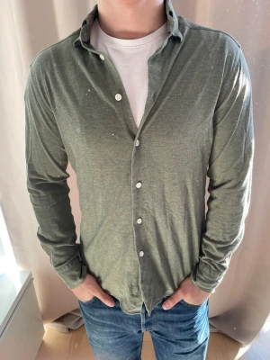 Grön långärmad skjorta med knappar - Säljer en stilren grön skjorta med klassisk krage och vita knappar. Skjortan är långärmad och har en normal passform, perfekt att bära över en t-shirt. Materialet känns mjukt och lätt, vilket gör den bekväm att ha på sig hela dagen.