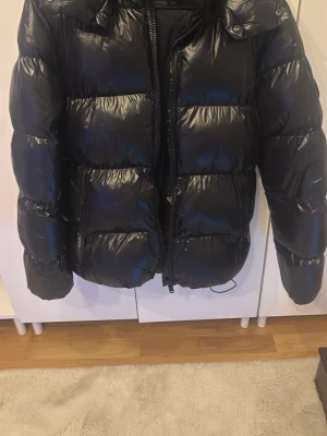 Svart glansig pufferjacka med huva - Säljer en svart, glansig pufferjacka med huva och dragkedja framtill. Jackan har stora, vadderade sektioner och två sidofickor. Perfekt för kalla vinterdagar och ger en riktigt trendig look.