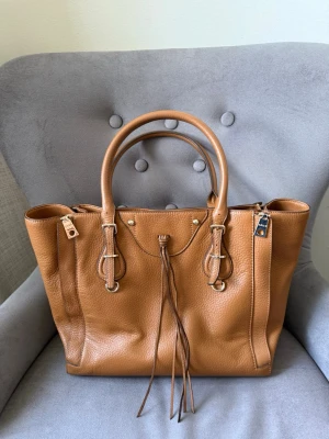 DKNY brun läderhandväska - Snygg handväska från DKNY i brunt, strukturerat läder med dubbla handtag och dekorativa läderdetaljer framtill. Väskan har guldiga metalldetaljer och en klassisk, rymlig form. Perfekt för dig som gillar stilrena och trendiga accessoarer.