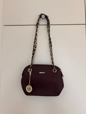 DKNY väska  - Söt liten cjute väska från DKNY, guldiga små detaljer lädret är i nån slags lila röd färg, blir skit snyggt till helsvarta outfits