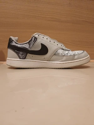 Nike vita och svarta låga sneakers - Nike sneakers i lågt utförande med vit bas och svarta detaljer. Skon har en klassisk siluett med svart swoosh på sidan och svart hälparti. Ovandelen är i läder och har perforeringar för ventilation. Snygg och tidlös modell för dig som gillar streetwear.