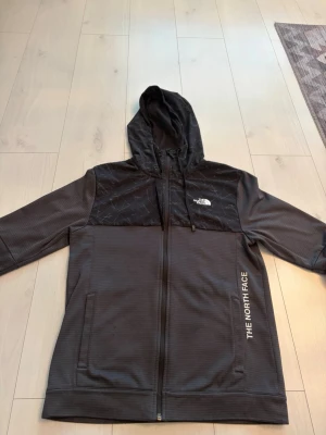 Svart hoodie från The North Face - Säljer en svart hoodie från The North Face med hel dragkedja och huva med snören. Överdelen har ett mönstrat tyg med logotyp på bröstet och text på sidan. Materialet är syntetiskt och känns sportigt och funktionellt. Perfekt för dig som gillar streetwear och outdoor.