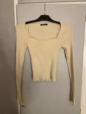  ribbad långärmad topp - Säljer en ljusbeige ribbad topp från Zara med lång ärm och snygg v-ringning. Toppen är slim fit och har en modern, croppad passform som framhäver siluetten. Perfekt att matcha med jeans eller kjol för en trendig look.