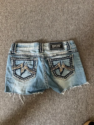 Blå Miss Me jeans med kontrastsömmar - Snygga blå jeans från Miss Me med tydliga vita kontrastsömmar och slitna detaljer. Klassisk femficksmodell med knappgylf och dekorativa sömmar längs fickor och midja. Perfekta för dig som gillar en unik denimlook.