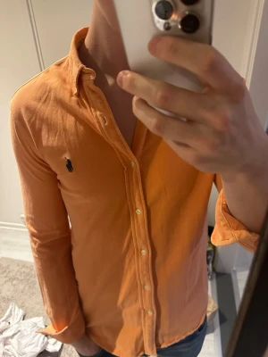 Orange skjorta från Ralph Lauren - Snygg orange skjorta från Ralph Lauren med klassisk button-down krage och broderad logga på bröstet. Skjortan har långa ärmar och vita knappar framtill. Perfekt för en stilren och avslappnad look.