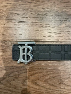 Svart Burberry bälte med silverdetalj - Jag kan gärna diskutera ett relativt rimligt pris. |Stilrent svart bälte från Burberry med klassiskt rutmönster och en stor silvrig B-spänne framtill. Bältet har en elegant och modern look som passar till många outfits. Perfekt accessoar för att lyfta din stil. 