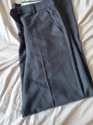 Mörkgrå kostymbyxor med raka ben - Snygga mörkgrå kostymbyxor med raka ben och klassisk passform. Byxorna har bälteshällor, snedställda fickor fram och är tillverkade i ett mjukt, vävt tyg som ger en stilren look. Perfekta för dig som vill ha en clean och tidlös stil.