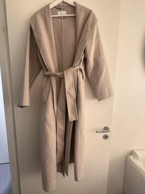 Beige ullkappa från Totême - Stilren beige ullkappa från STOCKHLM med knytskärp i midjan. Perfekt för dig som gillar minimalistisk och tidlös design. Nypris 1999kr. 