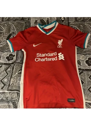 Liverpool matchtröja Nike röd äkta - Röd Liverpool FC matchtröja från Nike med klubbmärke och sponsortryck på bröstet. Tröjan har vita och turkosa detaljer vid krage och ärmslut samt vita sidor. Perfekt för fotbollsträning eller att bära på matchdag.