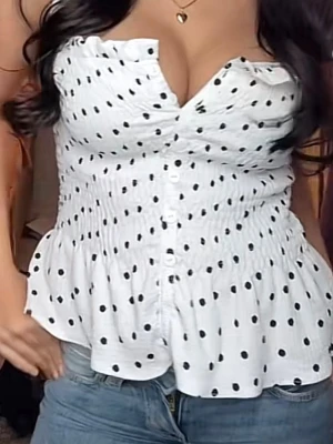 Vit prickig blus med volang - Supersöt vit blus med svarta prickar och volangkant nertill. Smockad överdel med knappar framtill och breda axelband med rynk. Perfekt för en trendig och somrig look.