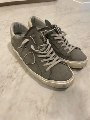 Philippe Model grå sneakers i mocka - Grå sneakers från Philippe Model i mjuk mocka med vita detaljer. Klassisk låg modell med grå skosnören och diskret logga på sidan. Snygg och stilren design som passar till det mesta. Nypris är 3000. Orginalbox följer med