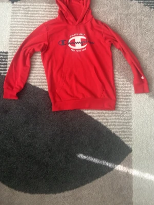 Röd Champion hoodie med logga - Röd hoodie från Champion med stor vit och svart logga samt text på bröstet. Klassisk känguruficka framtill och ribbade muddar vid ärmslut och midja. Mjuk bomullskänsla och huva för extra komfort. Perfekt för en sportig och avslappnad look.