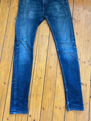 Replay jondrill jeans - Säljer ett par blå replay jeans med snygga slitningar och tvättade detaljer. Jeansen har klassisk femficksdesign och en liten reva på höger lår för en trendig look. Perfekta för dig som gillar en avslappnad men stilren stil. W29 L32