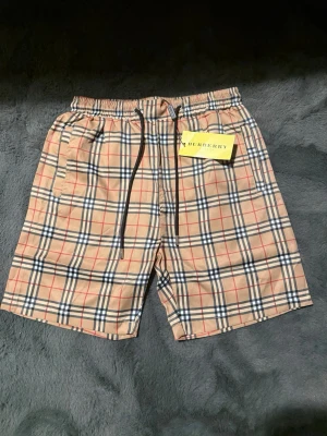 Rutig Burberry shorts med snörning - Säljer ett par klassiska Burberry shorts i beige med det ikoniska rutiga mönstret i svart, vitt och rött. Shortsen har elastisk midja med snörning och sidofickor. Perfekta för sommaren och riktigt snygga till en enkel t-shirt.