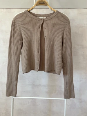 Beige ribbad kofta med knappar - Säljer en beige ribbad kofta med rund hals och långa ärmar. Koftan har en croppad passform och stängs med små knappar framtill. Perfekt att slänga över en topp för en chill look.