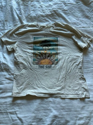 Vit t-shirt med tarotmotiv The Sun - Cool vit t-shirt med tryck av tarotkortet The Sun på bröstet. Motivet har sol, palmer och stjärnor i färgglada toner. T-shirten har rund hals och korta ärmar, perfekt för en avslappnad stil.