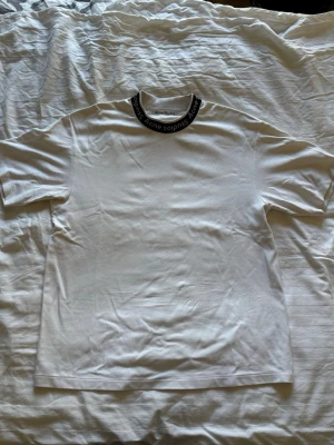 Vit t-shirt från Acne Studios - Vit t-shirt från Acne Studios med svart ribbad krage där varumärkets namn är tryckt runtom. Klassisk passform och korta ärmar. Tillverkad i mjuk bomull som känns skön mot huden. Perfekt för en clean och stilren look.