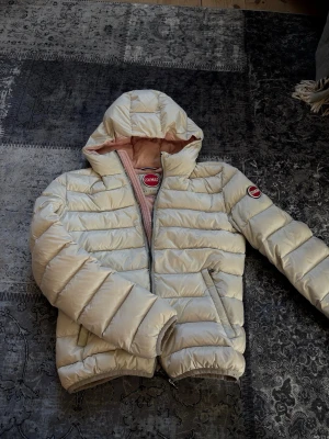 Beige jacka från Colmar - Snygg beige pufferjacka från Colmar med ljusrosa foder och huva. Jackan har en dragkedja framtill och Colmar-logga på ärmen. 