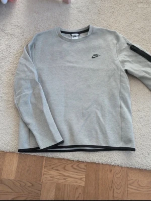 Grå nike tech tröja  kofta - Snygg grå sweatshirt från Nike med svart logga på bröstet och svarta detaljer vid hals, ärmslut och nederkant. Tröjan har rund halsringning och en dragkedjeficka på ena ärmen. Perfekt för en avslappnad och sportig stil. Har en jätte liten fläck i mitten men syns knappt