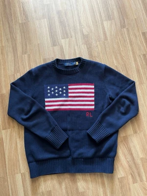 Mörkblå stickad tröja Polo Ralph Lauren - Mörkblå stickad tröja från Polo Ralph Lauren med amerikansk flagga i rött, vitt och blått på bröstet samt RL-broderi. Tröjan har rund halsringning och ribbade muddar vid ärmslut och nederkant. Perfekt för dig som gillar klassisk preppy stil.