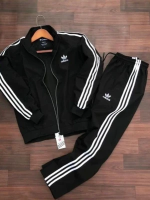 Svart Adidas träningsset med vita ränder - Snyggt svart träningsset från Adidas med klassiska vita ränder längs ärmar och ben. Setet består av en zip-jacka och matchande byxor, båda med Adidas logga på bröstet och benet. Perfekt för träning eller chill. Materialet är polyester.