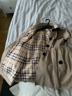 Beige trenchcoat med rutigt foder - Stilren beige trenchcoat med stora svarta knappar och klassiskt rutigt foder i beige, rött och svart. Jackan har markerad midja, fickor framtill och bred krage. Perfekt för dig som vill ha en tidlös och trendig look.