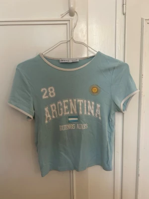 Ljusblå Argentina t-shirt Bershka - Ljusblå t-shirt från Bershka med vit kant vid hals och ärmar. Tryck med texten 'ARGENTINA', 'BUENOS AIRES', siffran 28, en liten sol och flagga på bröstet. Kortärmad och croppad modell, perfekt för en sportig och trendig look.