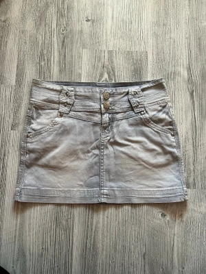 Ljusgrå kort jeanskjol med fickor - Snygg ljusgrå kort jeanskjol med dubbla knappar framtill, dekorativa sömmar och fickor både fram och bak. Kjolen har bälteshällor och silverfärgade nitar som detaljer. Perfekt för en trendig och avslappnad stil.(aldrig använd)