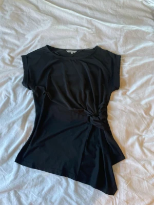 Svart asymmetrisk topp med knut - Svart topp med korta ärmar och snygg asymmetrisk nederkant. Toppen har en cool knutdetalj i sidan som ger en unik look. Tillverkad i mjukt och stretchigt material som sitter skönt på kroppen. Perfekt för dig som gillar stilrena och trendiga plagg.