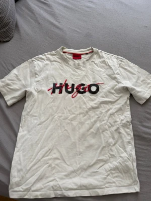 Vit Hugo Boss t-shirt med logga - Säljer en vit t-shirt från Hugo Boss med stor svart och röd logga på bröstet. Klassisk passform och rund halsringning. Perfekt för dig som gillar stilrena plagg med tydlig branding.