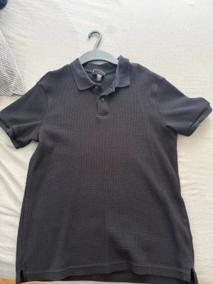 Svart pikétröja med krage - Klassisk svart pikétröja med korta ärmar och krage. Tröjan har två knappar vid halsen och är tillverkad i ett strukturerat bomullstyg som ger en snygg och enkel look. Perfekt till jeans eller shorts.