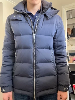 J.Lindeberg jacka marinblå - Snygg mörkblå dunjacka med huva och dragkedja framtill från J.lindeberg. Jackan har quiltad design, två sidofickor med dragkedja och hög krage. Perfekt för kalla vinterdagar och ger en stilren look. Den här många fina detaljer runt fickorna/huvan. Jätte snyggt J.lindeberg märke på ena armen. Jackan är sällsynt och säljs inte mer. Nypris är runt 5000kr.🤩🤩