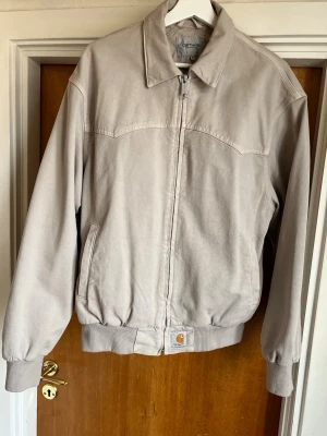 Beige Carhartt Jacka - Säljer en beige Carhartt jacka i modellen OG Santa Fe i Dusky Beige. Jackan har ribbade muddar vid ärmslut och midja samt klassisk krage. Materialet är slitstarkt bomullstyg och Carhartt-loggan sitter framtill vid nederkanten. Köpt ny i Carhartt butik, och anvendt bara nåra få gånger.