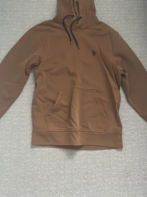 Brun polo hoodie med svart snöre - Snygg brun hoodie med huva och svarta dragsnören. Har en stor magficka framtill och en liten broderad logga på bröstet. Perfekt för en avslappnad och trendig stil. Mjuk bomullskänsla och ribbade muddar vid ärmslut och nederkant. Priset kan diskuteras (använd några gånger)