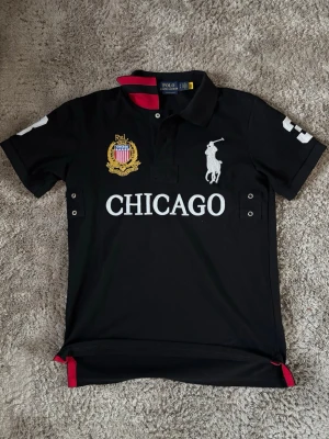 Svart Chicago polo från Polo Ralph Lauren - Snygg svart polotopp från Polo Ralph Lauren med stora vita broderade detaljer, CHICAGO-tryck och emblem på bröstet. Klassisk krage, korta ärmar och röda detaljer vid slitsarna. Perfekt för en sportig och stilren look.