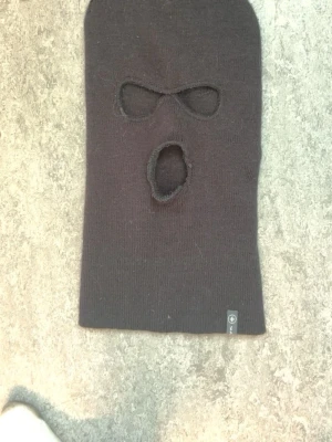 Svart balaclava mössa med hål - Svart balaclava mössa i stickat material med två hål för ögonen och ett för munnen. Märke C på liten tag vid nederkanten. Perfekt för kalla dagar när du vill täcka hela ansiktet. Enkel och stilren design.