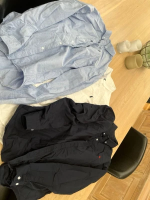 Ralph Lauren skjortor i bomull - Tre klassiska skjortor från Ralph Lauren i bomull. En ljusblå, en vit med broderad logga och en mörkblå. Alla har knappar framtill och lång ärm. Perfekta för en clean och stilren look.