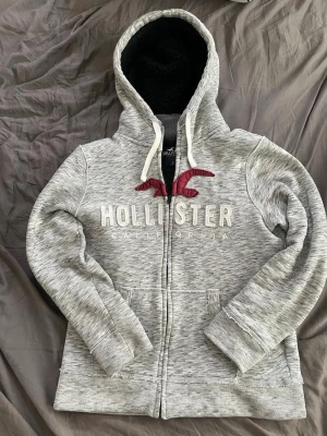 Grå Hollister hoodie med dragkedja - Snygg gråmelerad hoodie från Hollister med dragkedja framtill, stor huva med svart foder och vita snören. Broderad Hollister-logga och röd fågel på bröstet. Två fickor fram och ribbade muddar. Perfekt för chill dagar och streetwear vibes.