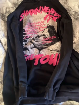 Shawn Mendes svart hoodie The Tour - Svart hoodie från Shawn Mendes med stor färgglad print på ryggen från The Tour. Trycket har rosa och silver detaljer samt ett porträtt av Shawn Mendes. Hoodien har huva och ribbade muddar. Perfekt för fans som vill sticka ut med snygg merch.